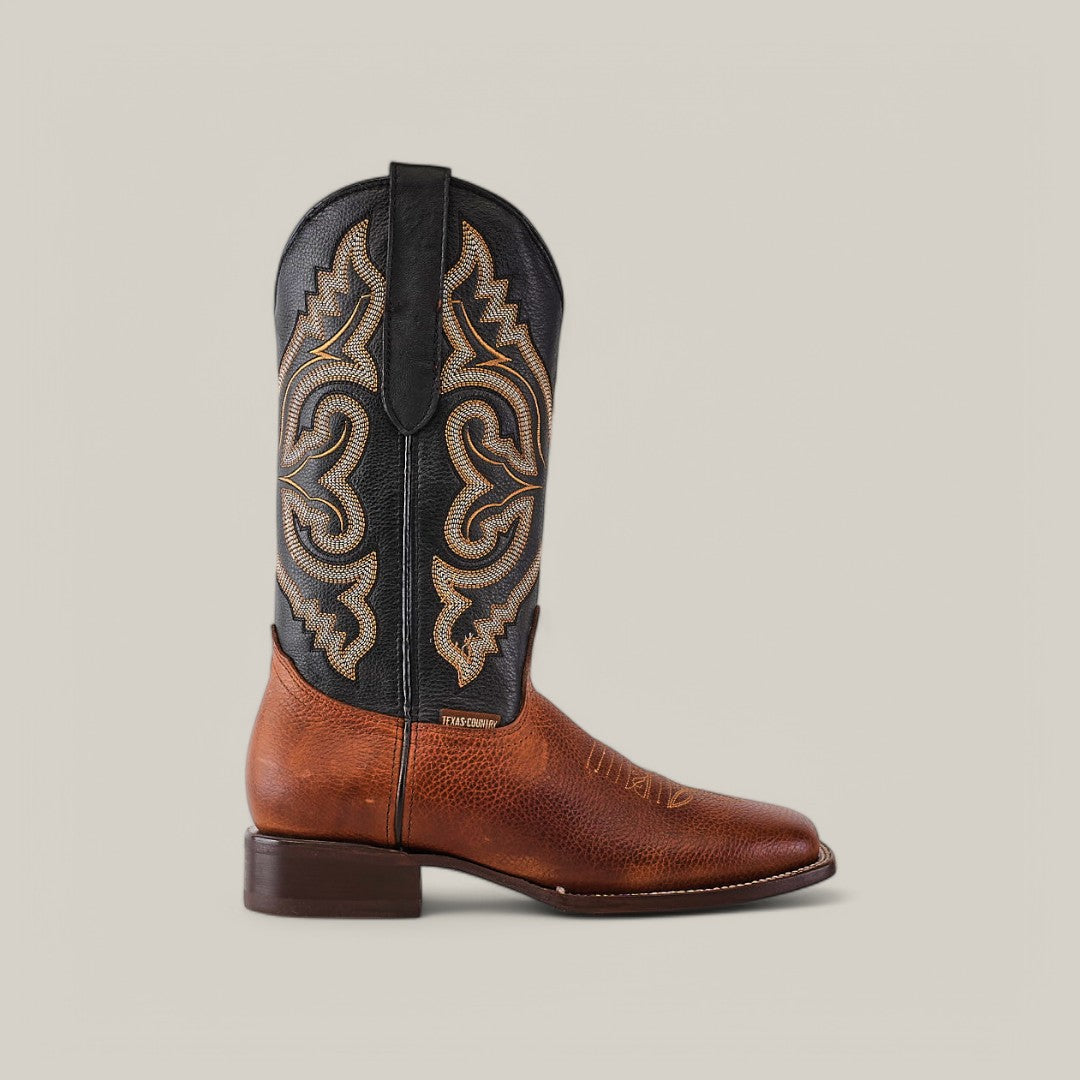 The Grisly Tan - Square Toe boot features Western elegance with a brown leather foot, a black shaft with intricate tan stitching, and a square toe with low heel, standing out against a plain light gray background.