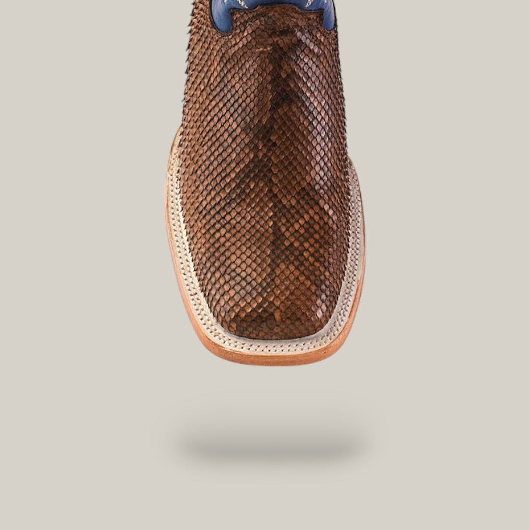 Close-up of the Exotic Python - Tamarindo shoe in brown python leather, tan sole, and blue accents atop its modern square toe design, set against a beige background where it appears to float with a soft shadow.