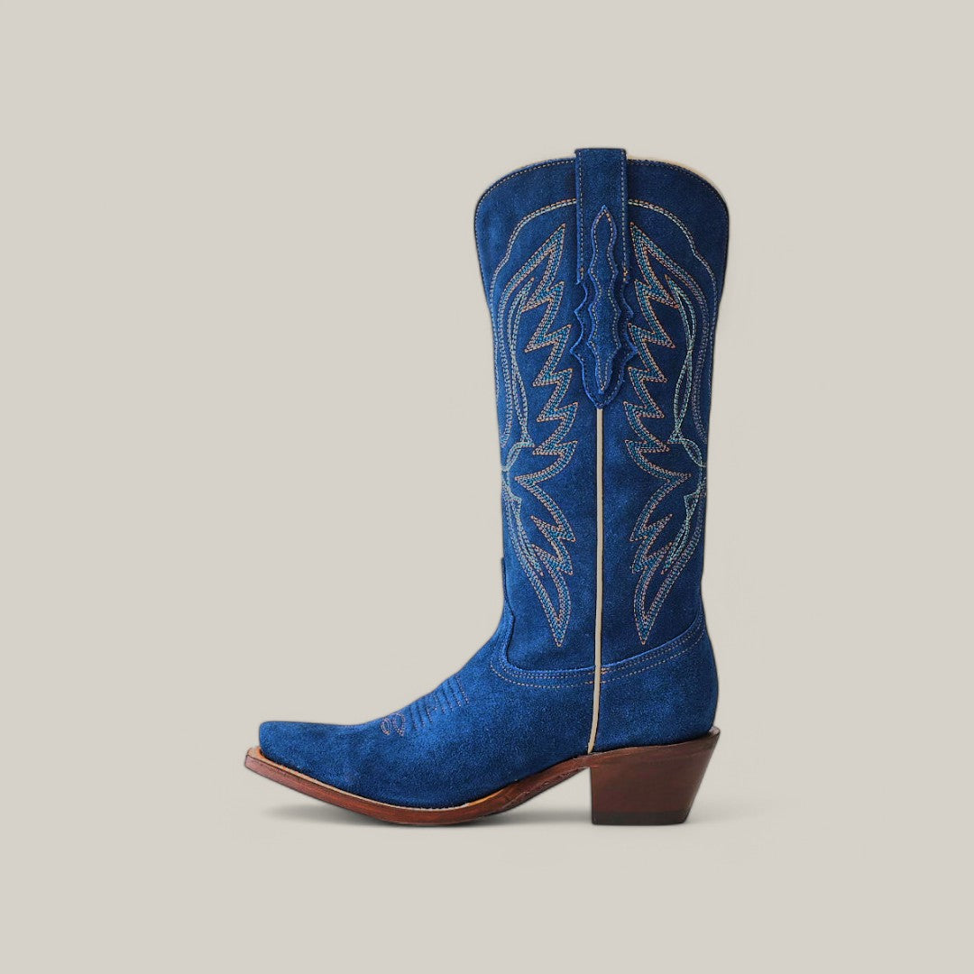 The Miranda Suede Blue Jeans boot features a mid-shaft design with intricate white stitching, set against a beige background. It showcases a snip toe, brown slightly angled heel, and durable leather sole.