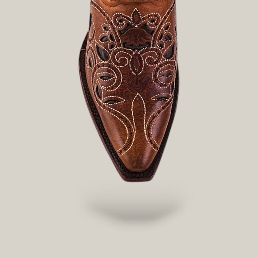 A close-up of the Gina - Brown - Snip Toe boot’s pointed toe showcases intricate floral stitching and cutout designs on brown premium leather against a light beige background.