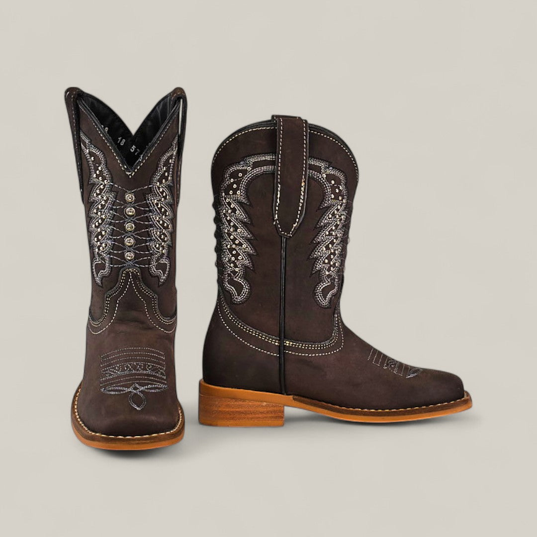 The Orion Suede Oxford - Square Toe Kids Boots feature brown suede with decorative white stitching and light brown soles. One boot stands upright while the other lies on its side, highlighting the patterned shaft and rounded toe design.