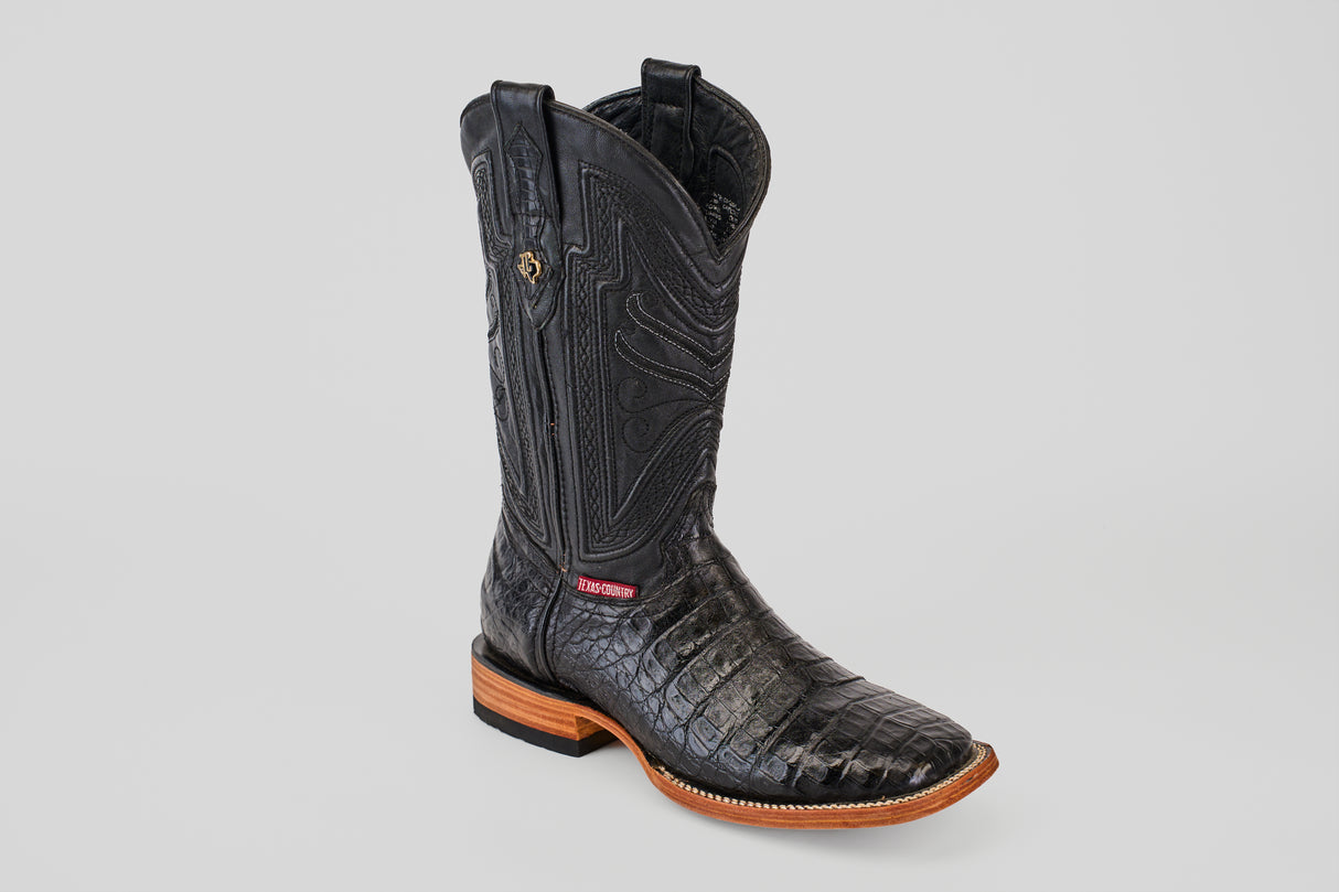 The Exotic Caiman Belly - Black- Square Toe boot features black caiman leather with an alligator texture, detailed shaft stitching, a brown leather sole, and a red ankle label, shown against a plain gray background.