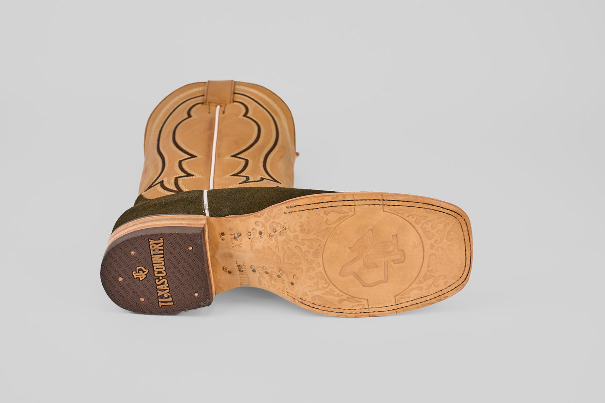 A green olive Prime Suede boot with a square toe lies on its side, displaying the sole with TEXAS COUNTRY branding, decorative patterns, and a stitched leather upper with curved designs.
