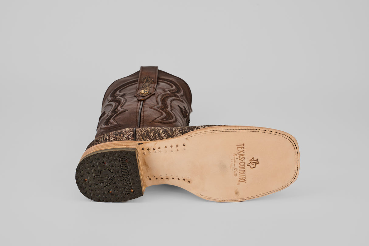 A brown Exotic Full Quilt Ostrich Leg - Matte Orix cowboy boot with a square toe lies on its side, showcasing the sole with “Texas Country” branding, tan leather, black heel, and decorative stitched details inspired by full quilt boots.