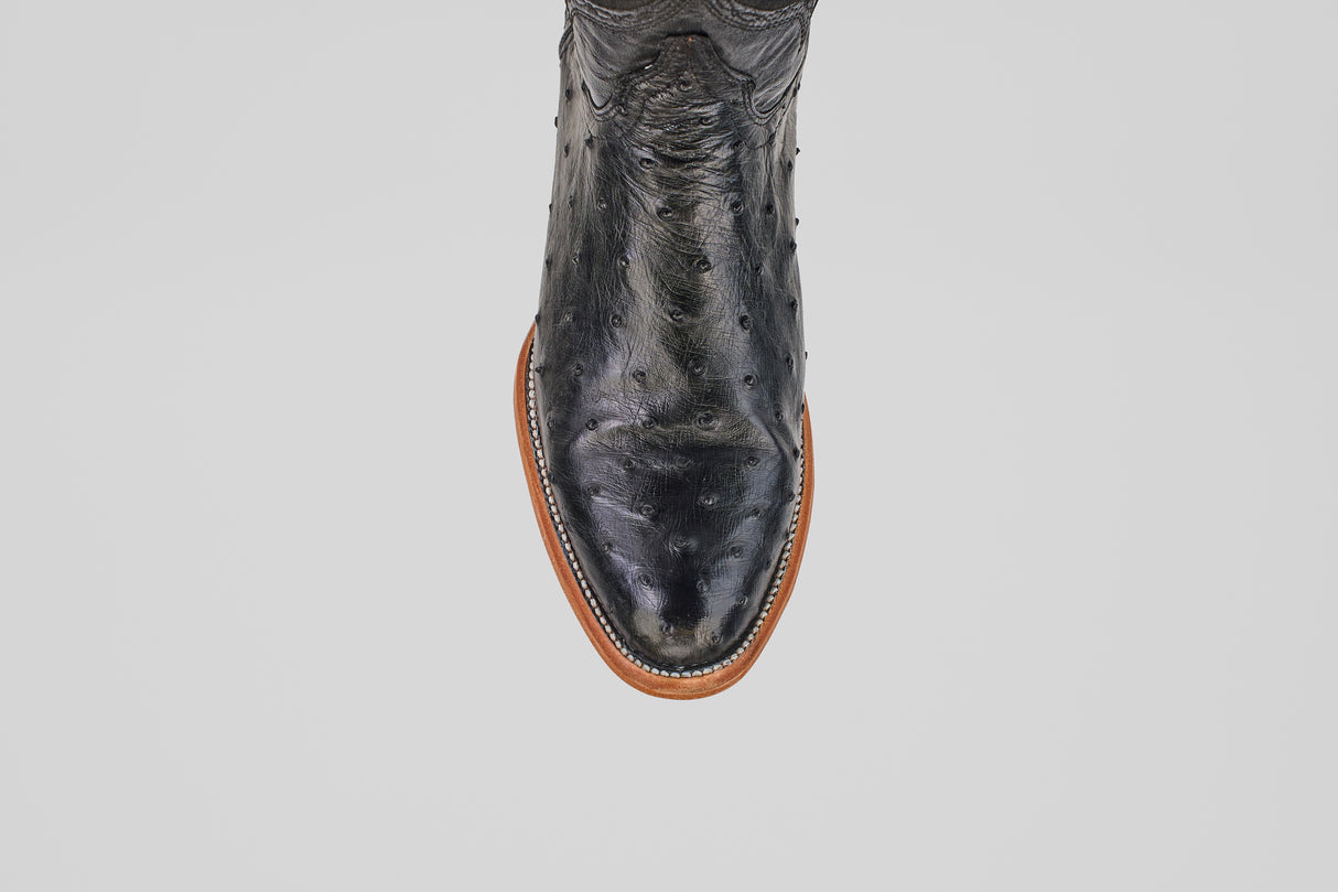 A close-up, top view of the Austin - Black - Roper Toe boot showcases its black textured ostrich leather and brown sole, highlighting refined western boot details against a plain white background.