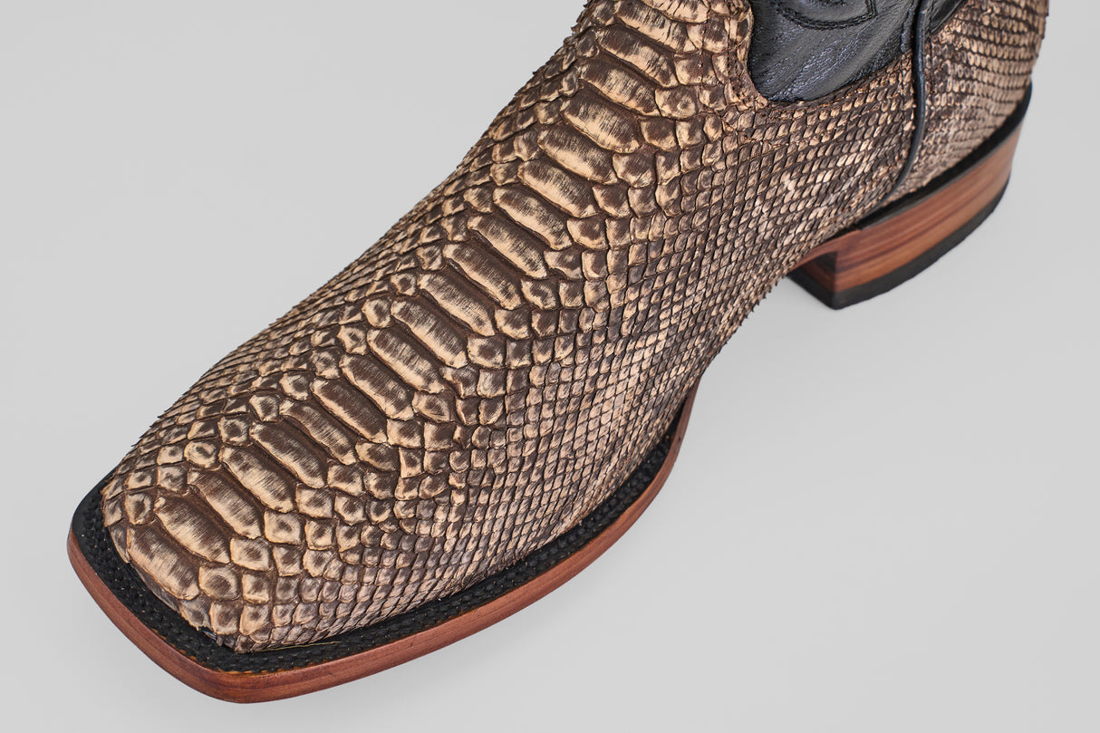 A close-up of the Tanner - Rustic Bone - Square Toe boot highlights its brown python design, black trim, and wooden sole, showcasing Western craftsmanship against a light gray background.
