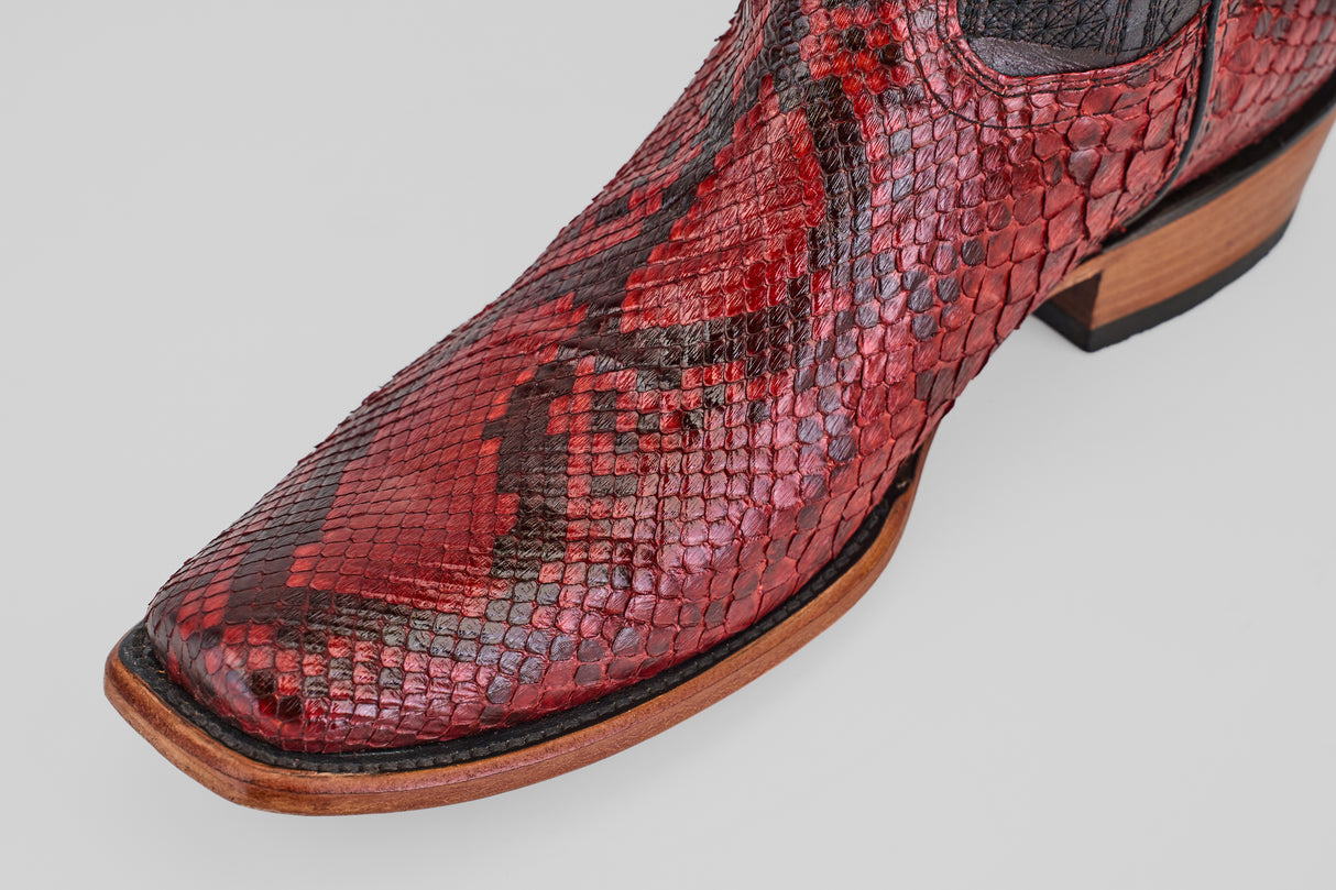 A close-up of the Tanner – Ruby – Narrow Square Toe boot, featuring red and black python leather, a wooden sole, and black stitching, set against a plain light gray background.