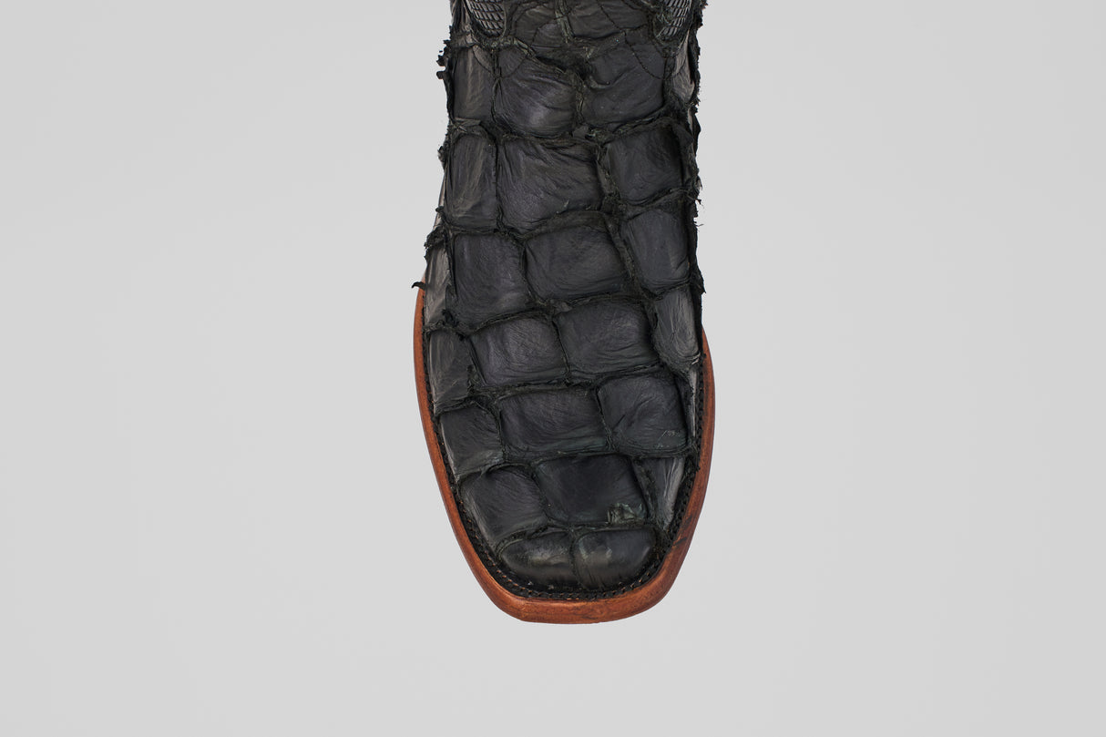 A close-up of the Conor - Matte Black - Narrow Square Toe boot shows its textured, fish leather-like pattern and brown sole, set against a plain gray background.