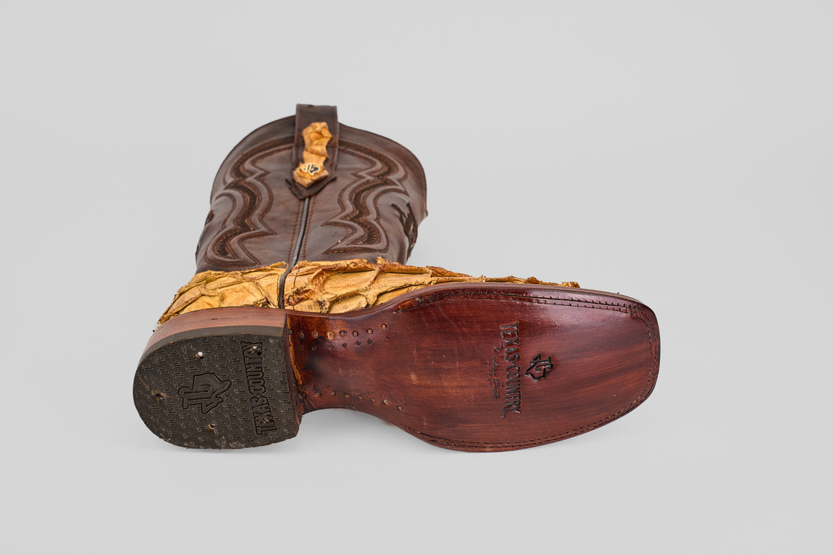 The Conor - Buttercup - Rodeo Toe boot, featuring brown leather with textured yellow detailing, lies on its side to reveal a dark wood sole engraved with text and symbols against a plain light background.