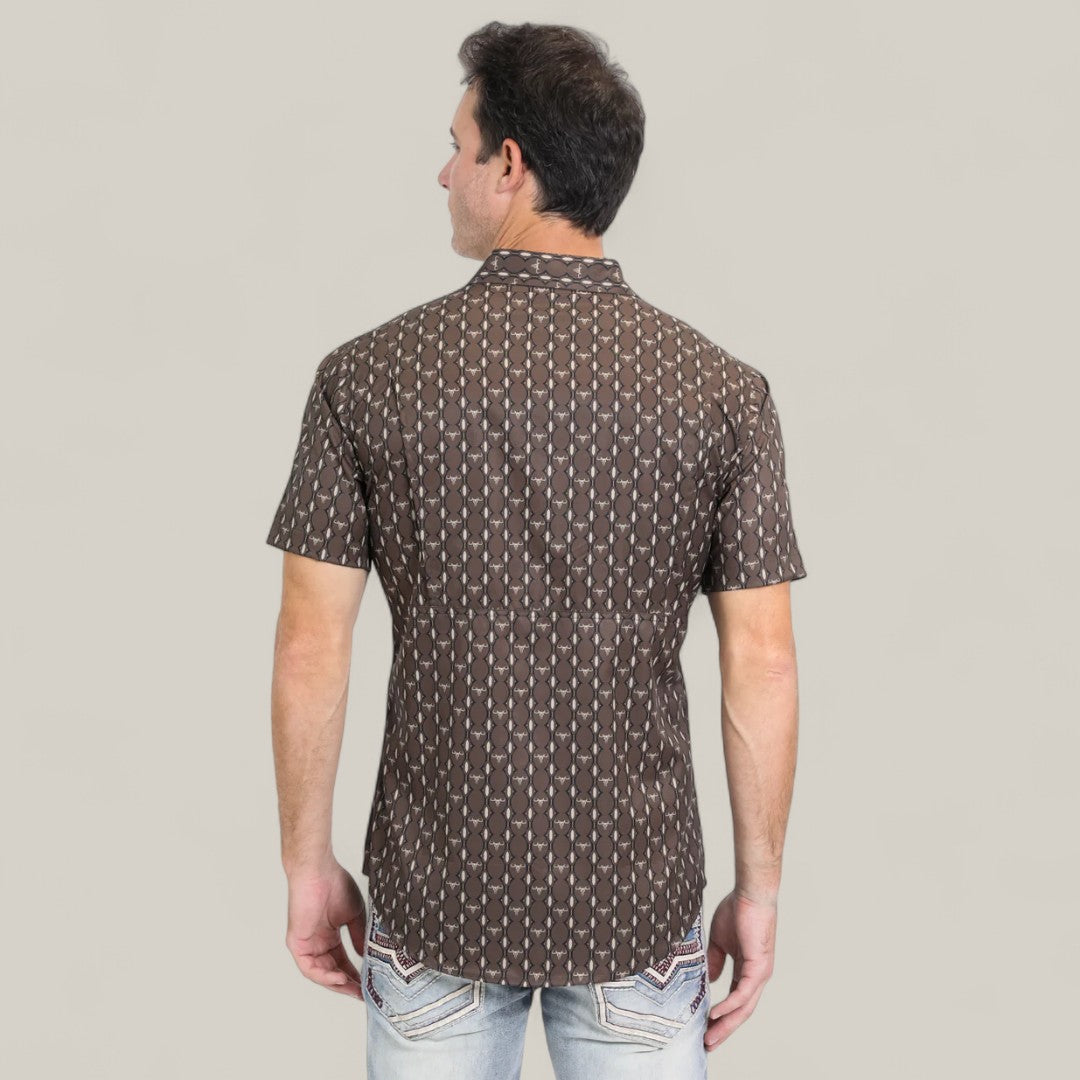 A man stands with his back to the camera, wearing a Mens Performance Western Short Sleeve Aztec Print Brown Shirt - PGW10643 and light blue jeans with decorative pocket stitching, set against a plain light background.