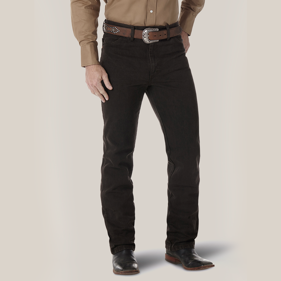 A person wearing a tan long-sleeve shirt, Wrangler Cowboy Cut Slim Fit Jean (0936KCL), a studded brown belt with a large buckle, and black boots. The backdrop is plain white.