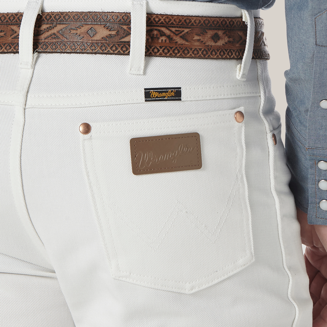 A person wears Wrangler Cowboy Cut Slim Fit White Jeans - 0936WHI, complemented by a brown leather belt. The jeans feature a Wrangler patch and back pocket label. A chambray shirt with patterned snaps adds rugged sophistication.