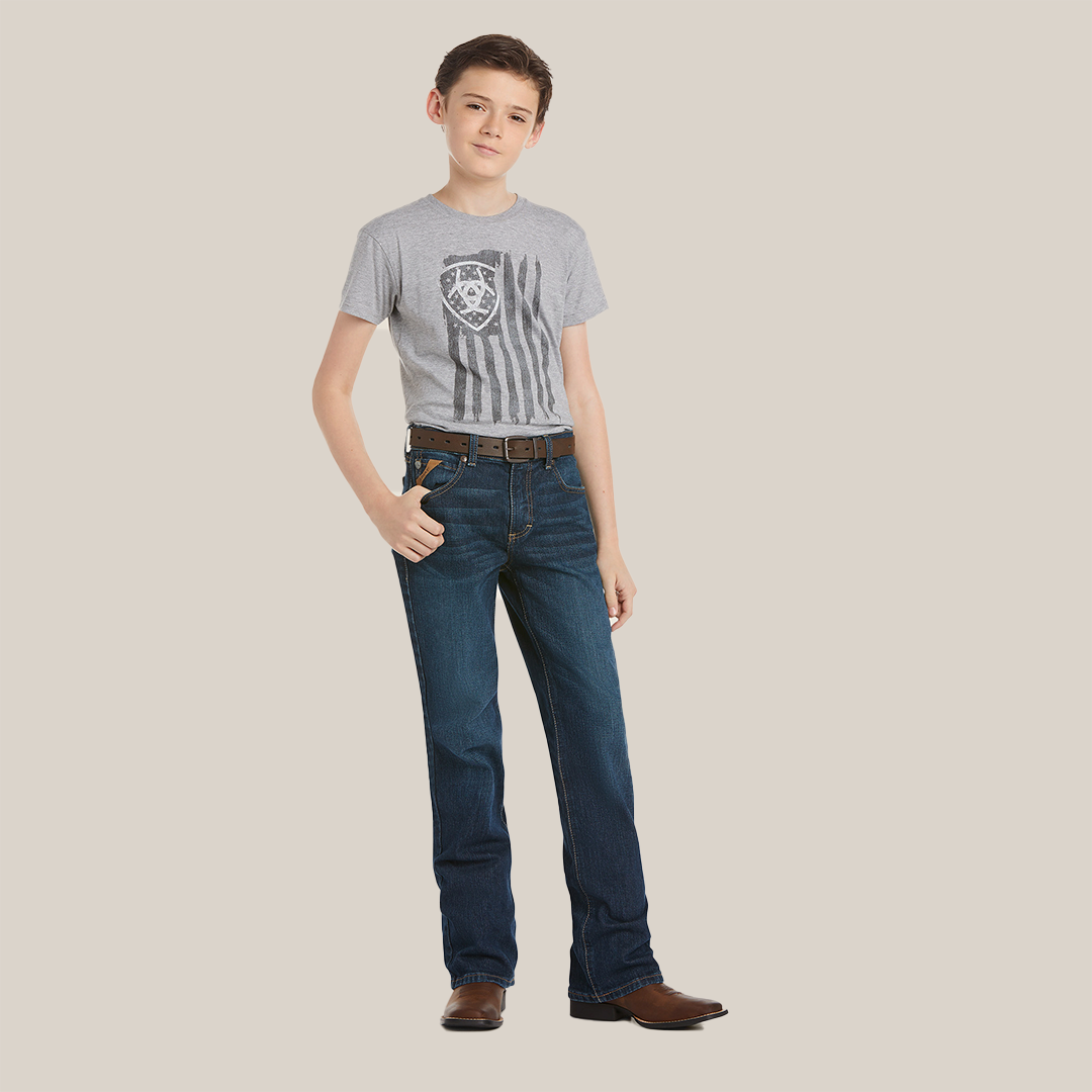A young person wearing a gray T-shirt with a shield and stripes design, Ariat Boys B4 Legacy Dark Wash Relaxed Fit Boot Cut Jeans, and brown cowboy boots stands confidently with one hand in their pocket against a neutral background.