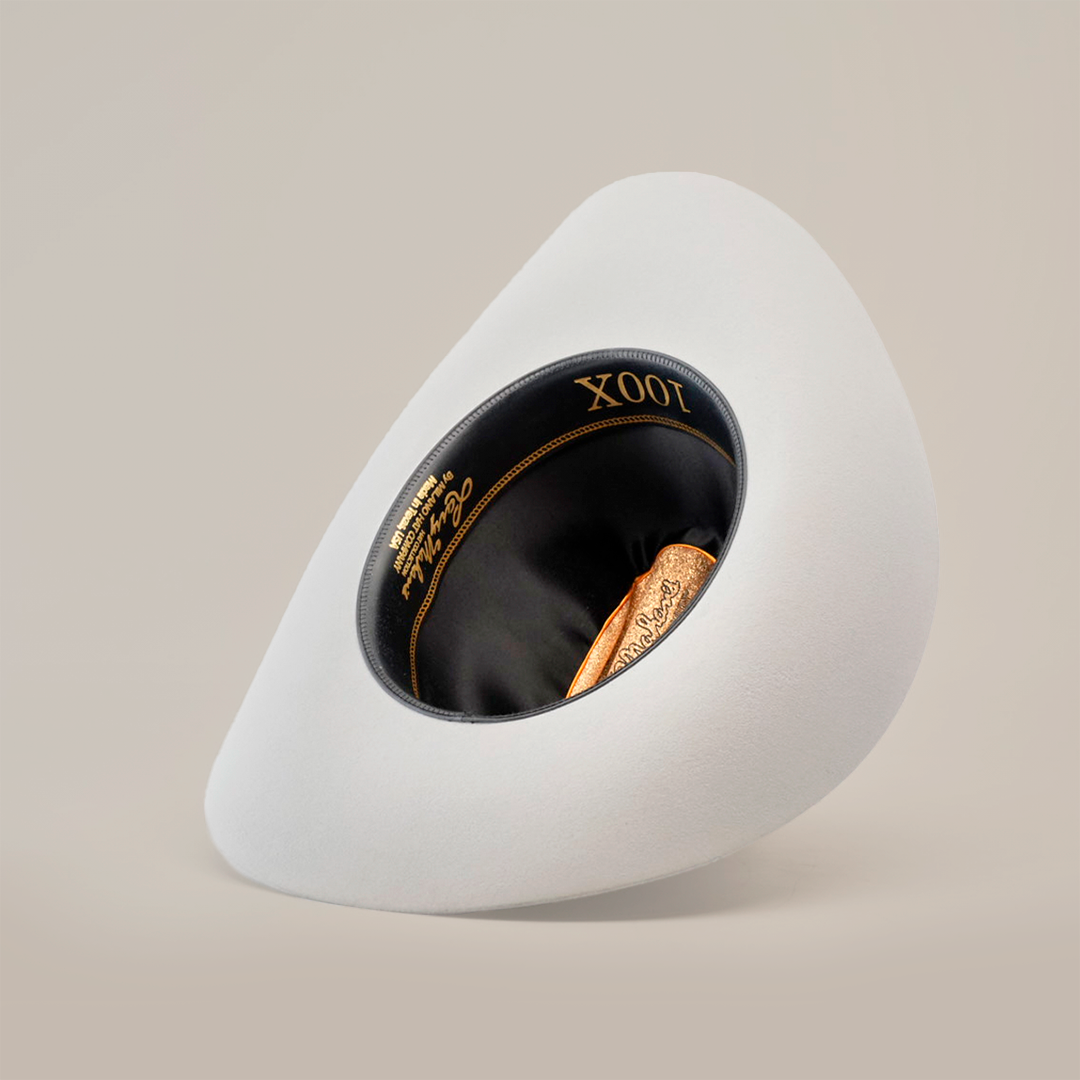 The 100X Independecia Platinum, a white asymmetrical oval-shaped hat epitomizing Western headwear, features a black interior with Larry Mahan 100X in gold script inside the brim, elegantly tilting on a plain, light background in El Presidente style.