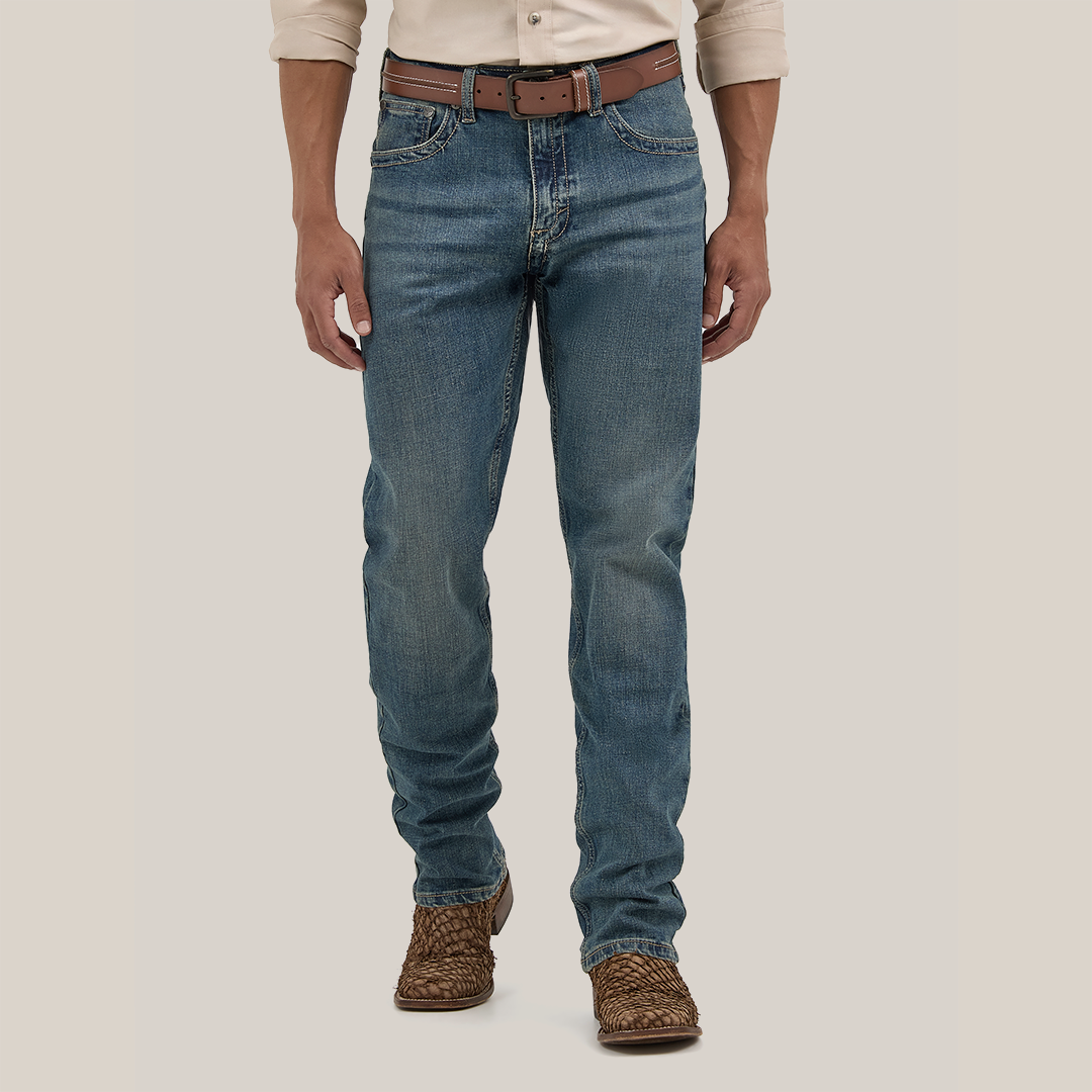 The lower half of a person is visible, wearing Wrangler Mens 20X Extreme Relaxed Fit Jeans - 1044MWXMA in Premium Denim blue with a worn look. The outfit is completed with a beige shirt, brown leather belt, and woven brown shoes against a plain background.