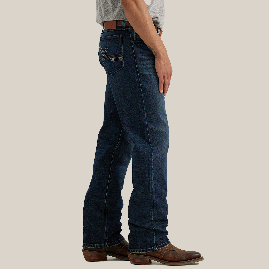 A man in a gray t-shirt and leather belt stands sideways wearing dark blue Wrangler 20X Sunnybrook Dark Wash Extreme Relaxed Straight Stretch Denim Jeans, featuring visible back pocket stitching, and brown cowboy boots. The background is plain gray.