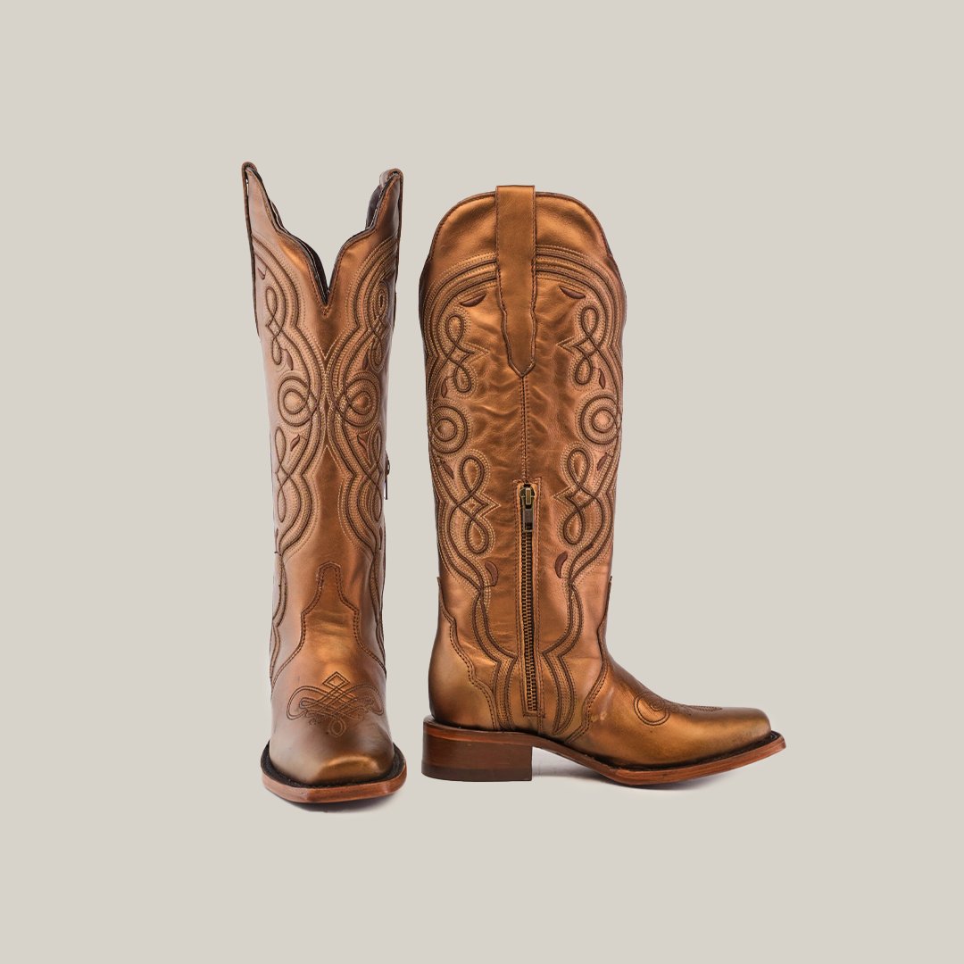 A pair of brown cowboy boots with intricate leather stitching are positioned against a plain neutral background. One boot stands upright while the other is slightly tilted, showcasing the side zipper.