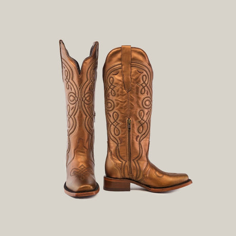 A pair of brown cowboy boots with intricate leather stitching are positioned against a plain neutral background. One boot stands upright while the other is slightly tilted, showcasing the side zipper.