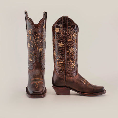 A pair of dark brown cowboy boots with intricate gold embroidery featuring floral and swirl patterns, standing on a light background.