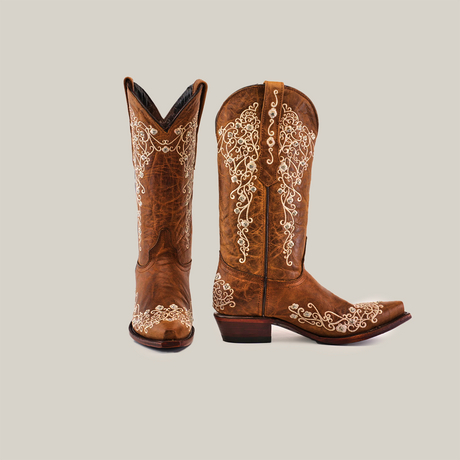 A pair of brown cowboy boots with intricate white embroidery. One boot is upright while the other is angled slightly to the side, showing the detailed design on both sides. The background is a neutral color.