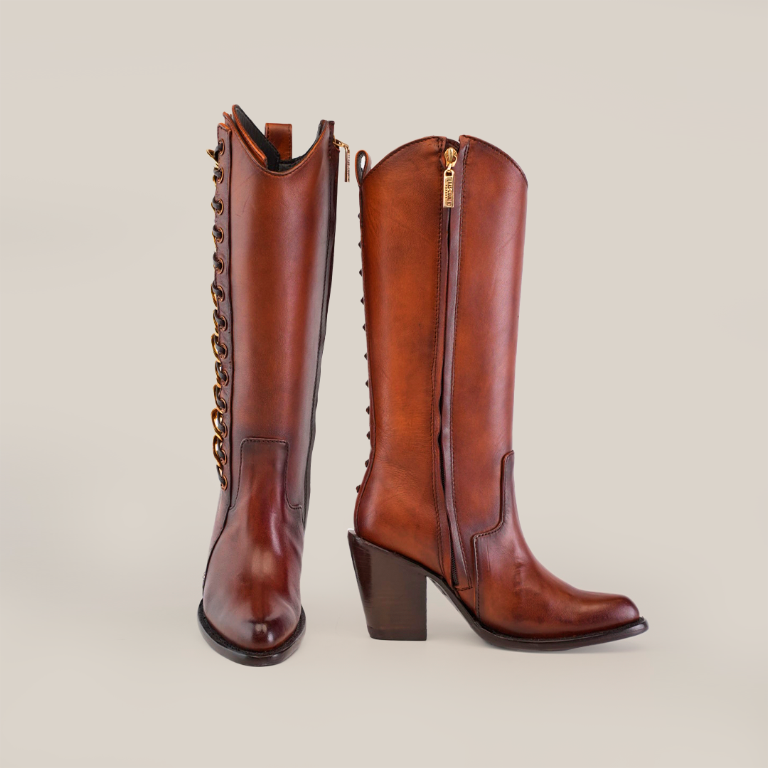 A pair of brown leather cowboy boots with block heels, gold zippers on the side, and gold chain detailing on the back, set against a plain beige background.