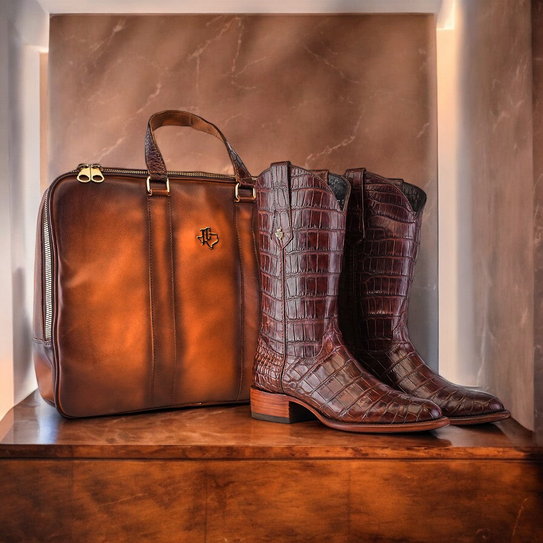 A pair of brown crocodile-patterned cowboy boots stand next to a brown leather briefcase on a wooden shelf, against a marble-patterned wall.