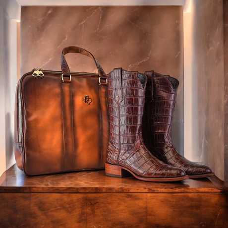 A pair of brown crocodile-patterned cowboy boots stand next to a brown leather briefcase on a wooden shelf, against a marble-patterned wall.