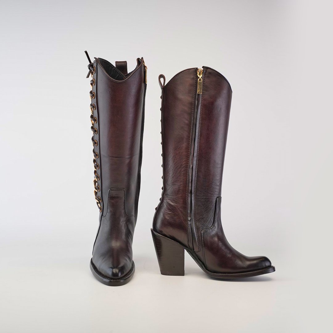 Two brown leather cowboy boots with block heels and sleek design. One boot shows a front view with decorative laces, and the other shows a side view with a visible zipper. Background is light gray.