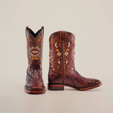 A pair of brown cowboy boots with intricate orange, yellow, and white geometric patterns on the shafts, displayed against a plain background. One boot stands upright, and the other lies on its side.