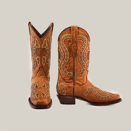 A pair of ornate brown cowboy boots, one upright and one slightly tilted, featuring intricate cutout designs and detailed embroidery on a plain background.