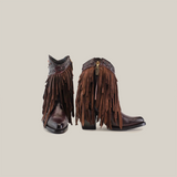 The Austin Fringe Bootie Chocolate J Toe exudes Western charm with its brown leather, layered fringe details, decorative band with a circular pattern, golden pull tab on the side zipper, pointed toe, and chunky heel.