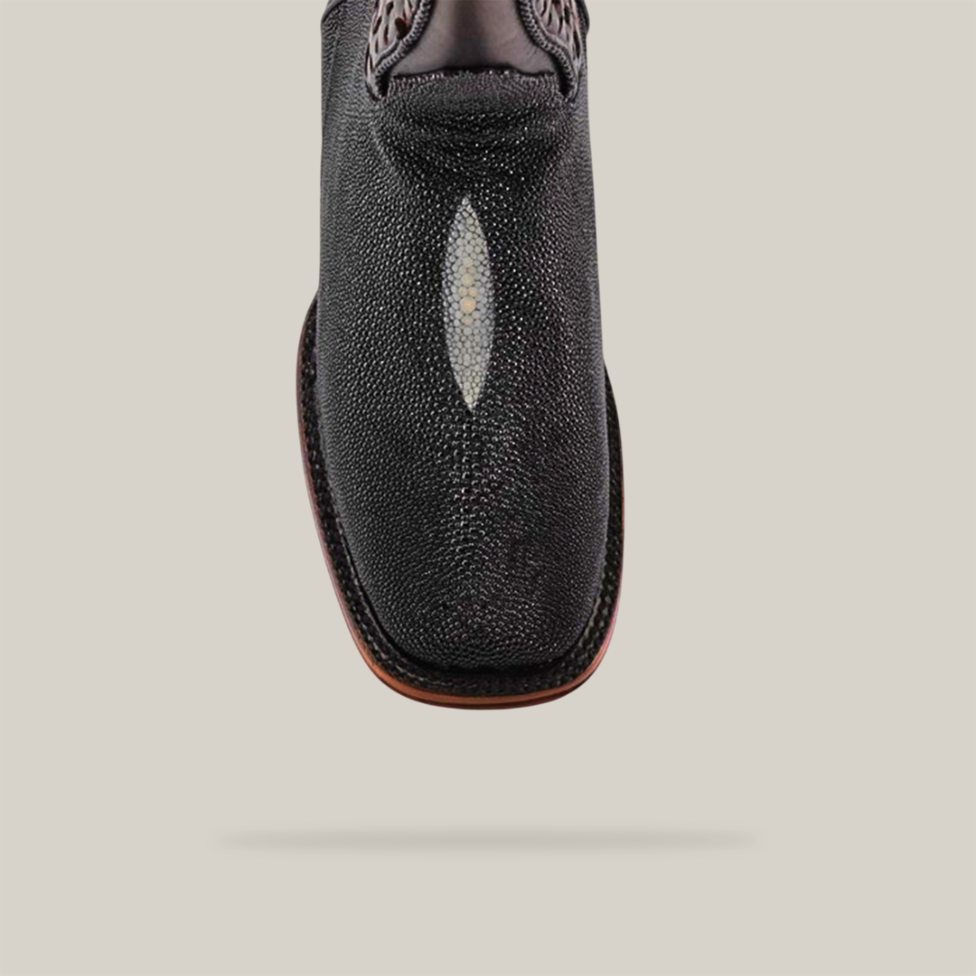 A detailed view of the Exotic Stingray - Herradura black cowboy boot shows its handcrafted quality and intricate stitching. It has a decorative white oval and a square toe, highlighted against a plain white background. The rough texture hints at the durability of its Goodyear welt sole.