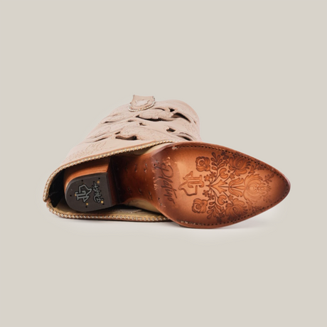 The Roma Flowers Tall Shaft Cream - J Toe boot is shown from the bottom, highlighting intricate floral engravings on the leather sole. It features decorative stitching and a pointed toe, all set against a plain white background.