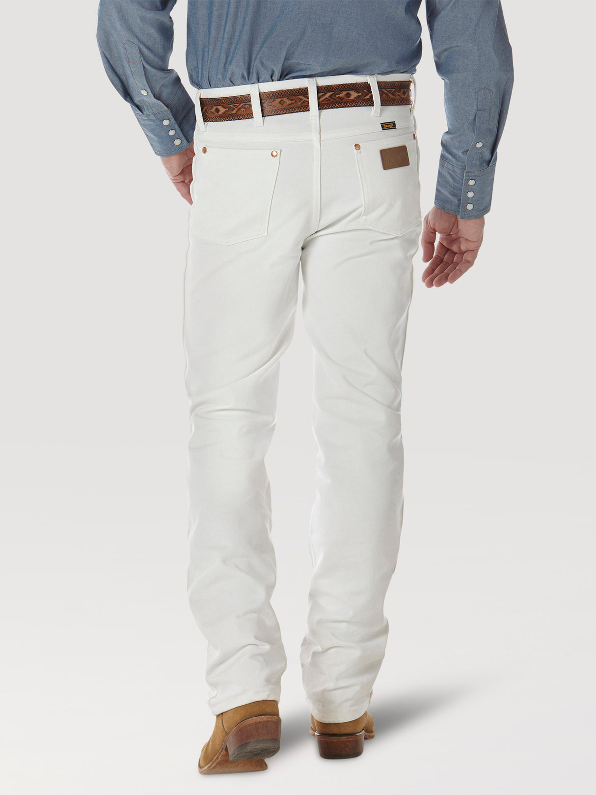 Against a light backdrop, someone is wearing Wrangler Cowboy Cut Slim Fit Jean in white, paired with a blue long-sleeve shirt, brown shoes, and a patterned brown belt. The ensemble subtly suggests a classic cowboy style.