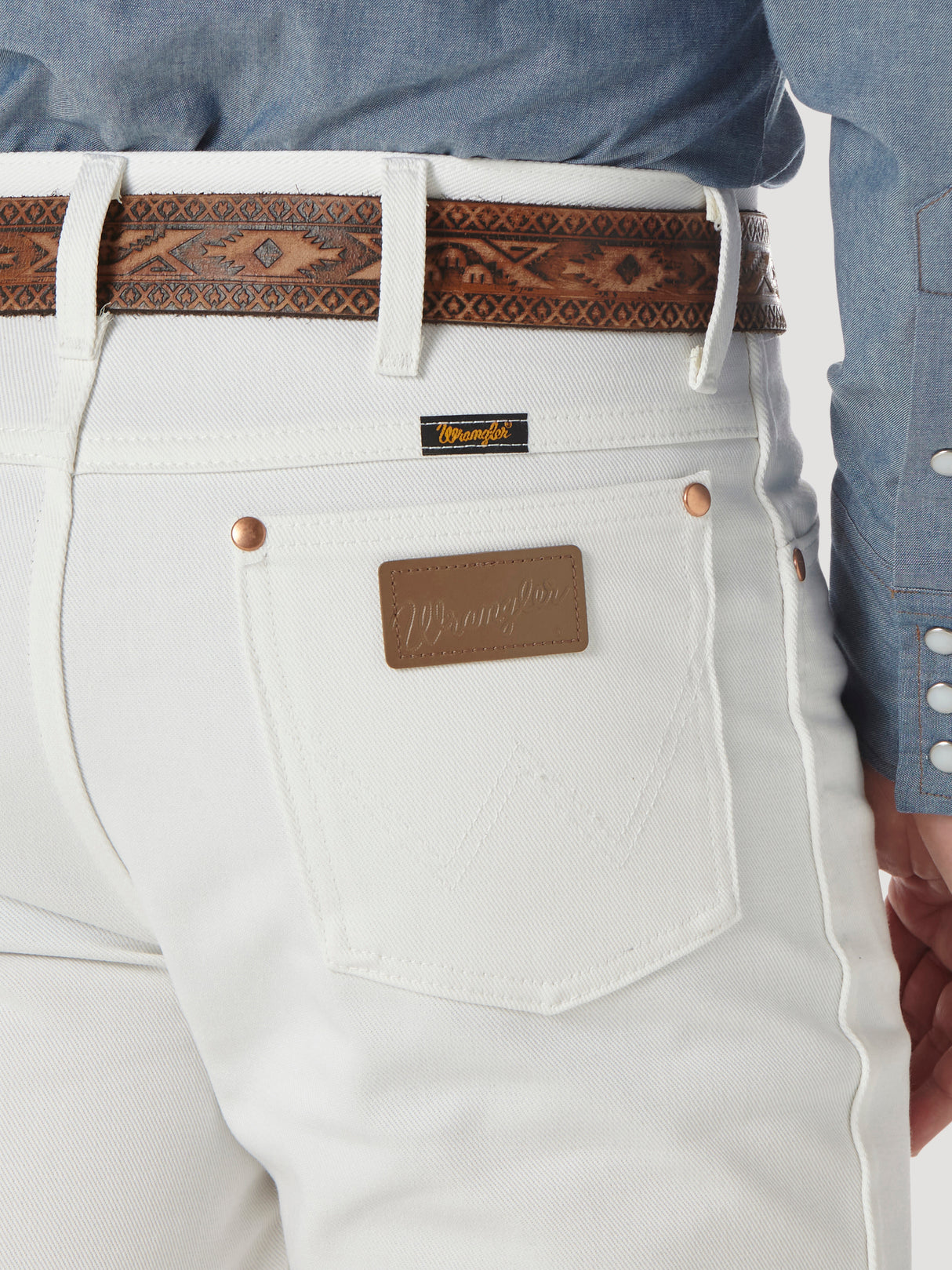 A person wears Wrangler Cowboy Cut Slim Fit White Jeans - 0936WHI, complemented by a brown leather belt. The jeans feature a Wrangler patch and back pocket label. A chambray shirt with patterned snaps adds rugged sophistication.