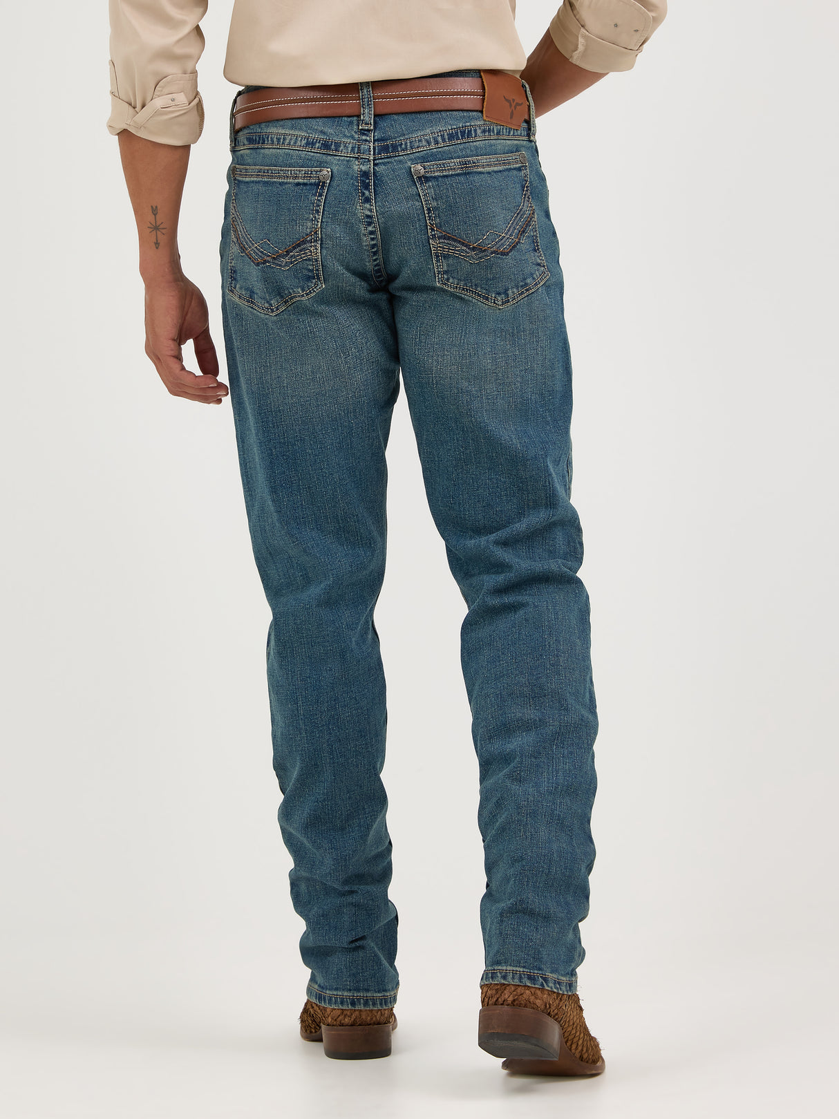 A person in Wrangler Mens 20X Extreme Relaxed Fit Jeans - 1044MWXMA, blue denim with a brown belt and shoes, stands against a white background. They sport an arm tattoo and light-colored rolled sleeves, epitomizing the relaxed style.