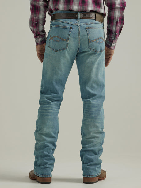 A person in Wrangler 20X Mens Light Wash Shade Bootcut Stretch Jeans and a plaid shirt stands facing away. The premium denim jeans boast decorative stitching on the back pockets, complemented by a brown belt and shoes, set against a plain, neutral backdrop.