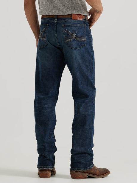 A person in a gray shirt and Wrangler 20X Sunnybrook Dark Wash Extreme Relaxed Straight Stretch Denim Jeans with decorative back pocket stitching stands on a plain white background. They complement the look with brown leather boots and a matching belt, highlighting the jeans relaxed fit.