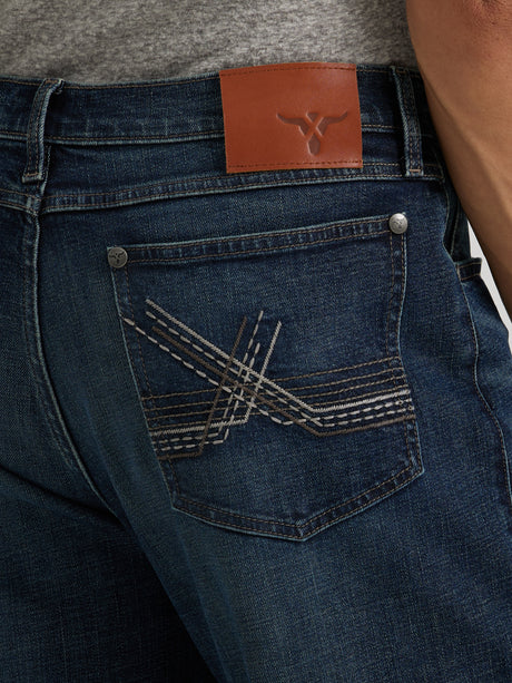 A person wears Wrangler 20X Sunnybrook Dark Wash Extreme Relaxed Straight Stretch Denim Jeans, paired with a gray shirt. The premium jeans feature decorative stitching on the back pocket and a brown leather patch with a small logo on the waistband.