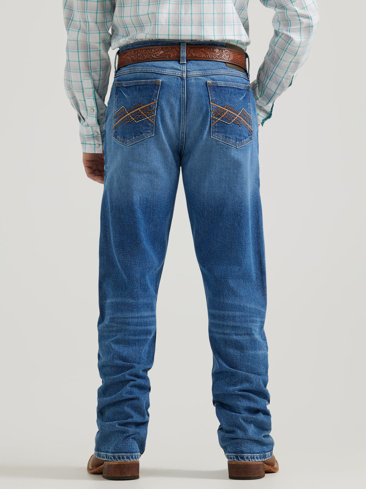 A person stands facing away wearing Wrangler 20x Mens Medium Wash Backwater Vintage Bootcut Stretch Denim Jeans with decorative stitching, a plaid shirt, and brown leather sandals against a light gray background.