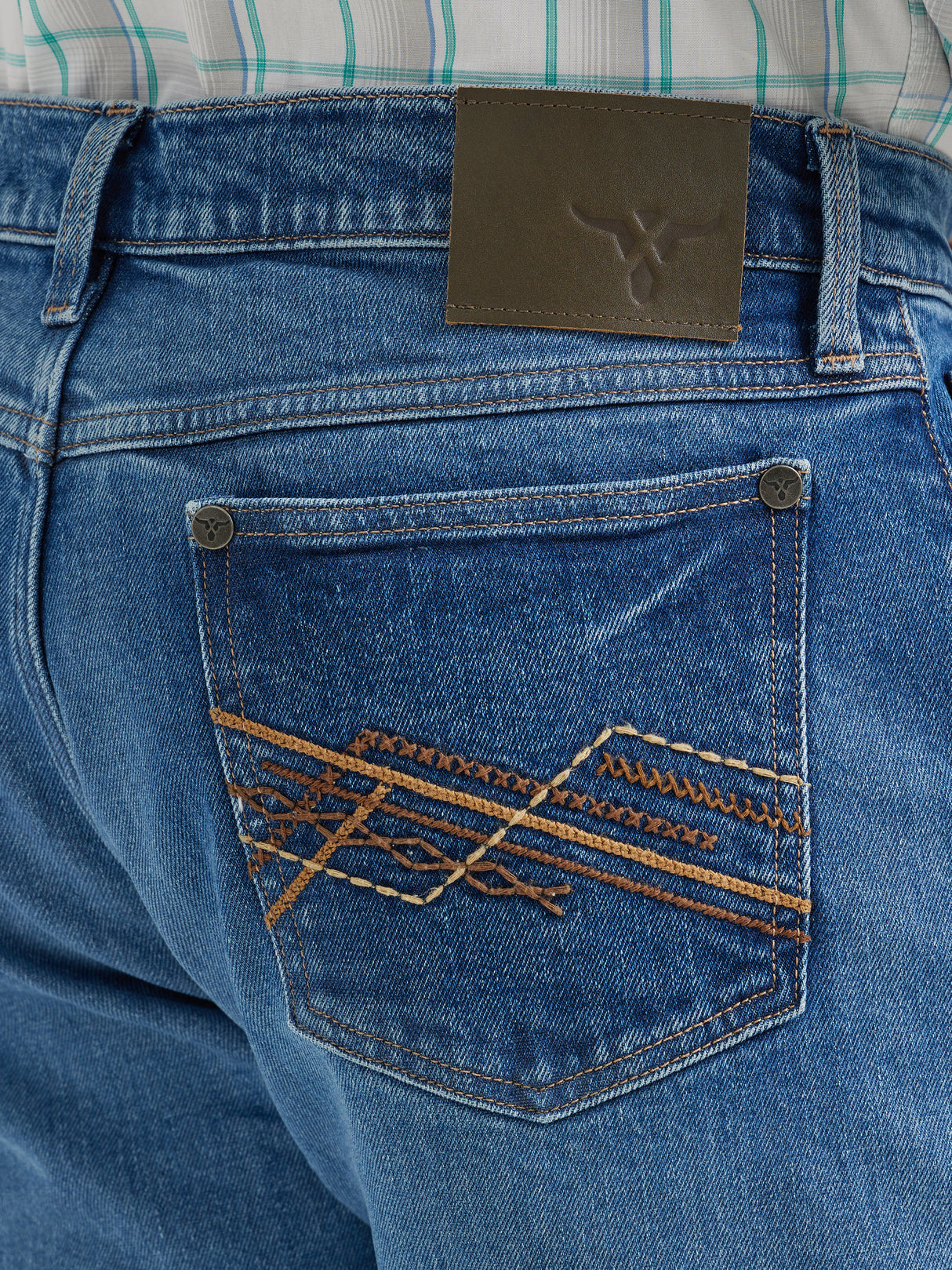 A close-up shows the back pocket of Wrangler 20x Mens Medium Wash Backwater Vintage Stretch Jeans, featuring an orange and brown embroidered geometric design. A logo leather patch is on the waistband. The jeans pair classically with a tucked plaid shirt for a timeless style.