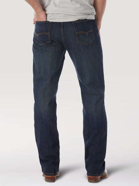 A person poses against a white backdrop wearing the Wrangler 20X No. 33 Extreme Appleby Dark Wash Relaxed Straight Leg Jeans, featuring orange stitching and styled with brown shoes, embodying a laid-back essence with hands in back pockets and a light gray shirt.