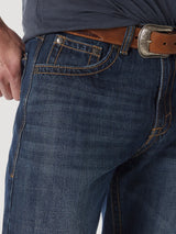 A person is wearing Wrangler® 20X® No. 33 Relaxed Straight Leg 1033MWXWL jeans with orange stitching and a brown belt with a silver buckle. The right hand is partially visible, resting near the front pocket of the relaxed fit jeans.