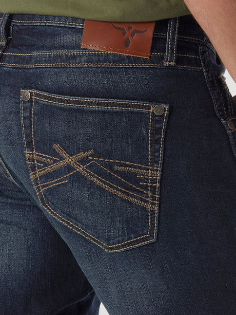 A close-up shows a person in Mens Wrangler 20X No. 42 Vintage Bootcut Jeans. These dark blue slim fit jeans boast tan stitching with intricate patterns on the right back pocket and a brown leather logo patch on the waistband, perfectly complementing a green shirt.