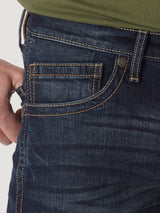 A person wearing Mens Wrangler 20X No. 42 Vintage Bootcut Jean - 42MWXRD in premium dark blue denim, accented with orange stitching. With a hand adorned by a silver ring tucked into the pocket and paired with a green shirt, the stylish slim fit jeans exude comfort.