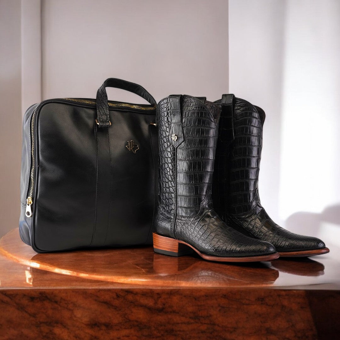 An Exotic Full American Alligator - Black - Round Toe cowboy boot with brown soles sits beside a luxury briefcase on a polished wooden surface, against a softly lit white wall.