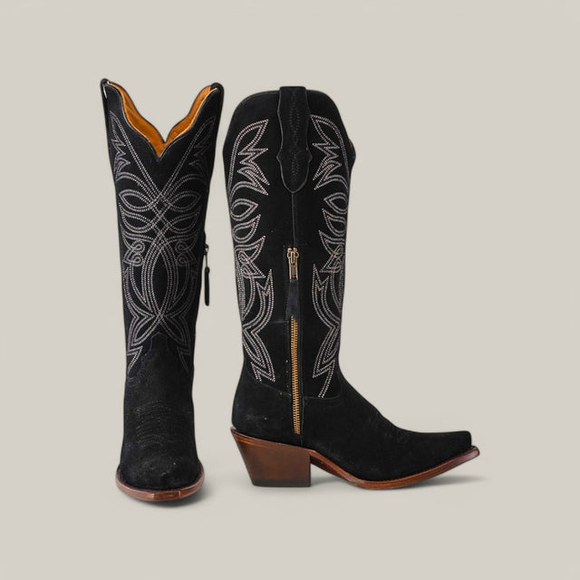 The Kellie Suede Black Leather Sole boots feature intricate white stitching and a snip toe. The left boot is upright, slightly turned to display its side zipper, while the right is angled to showcase its heel and pointed toe with a brown finish.