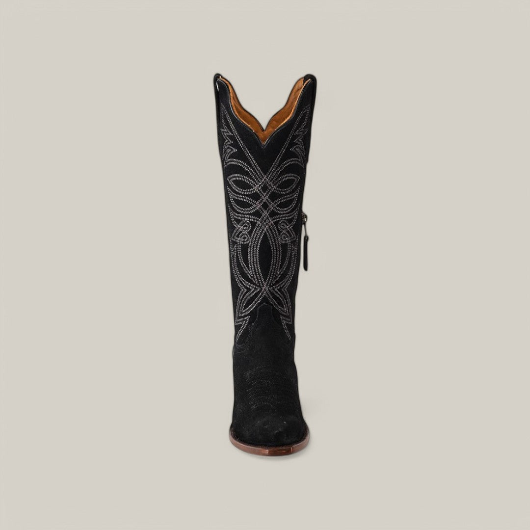 The Kellie Suede Black boot showcases intricate white embroidery on its tall shaft, set against a light gray background. The design features swirling patterns, complemented by a brown leather sole and snip toe.