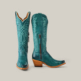 The Kellie Turquoise Leather Sole boots feature intricate white stitching, a snip toe, black side zippers, and a wooden heel on tan soles. Set against a plain gray background, these tall shaft boots stand out with their unique design.