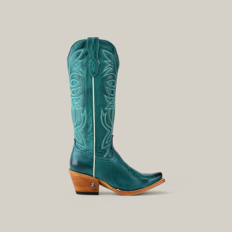 The Kellie Turquoise Leather Sole boot, featuring a tall shaft and snip toe, showcases intricate stitching. Its wooden stacked heel enhances the polished leathers vibrant look against a light beige background.